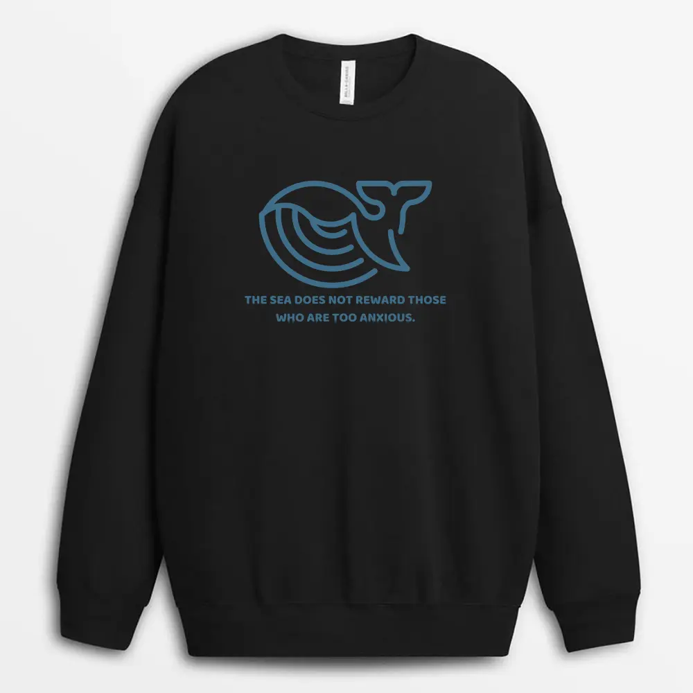Model seated on a rocky coastline wearing a charcoal sweatshirt with dramatic sea imagery on the back, conveying endurance and inner challenge.